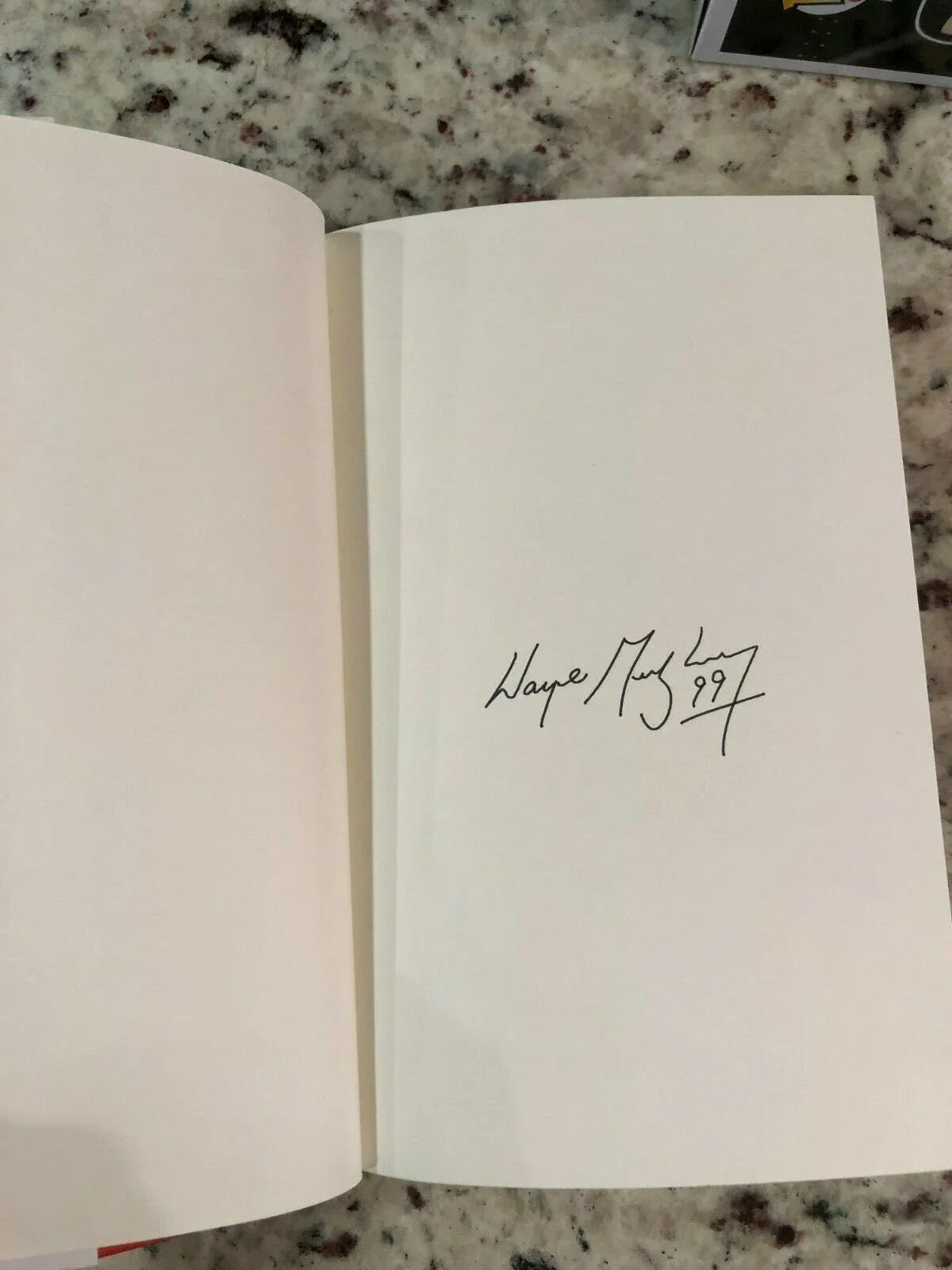 Open book showing a black ink signature with date '99' on a blank page against a granite tabletop